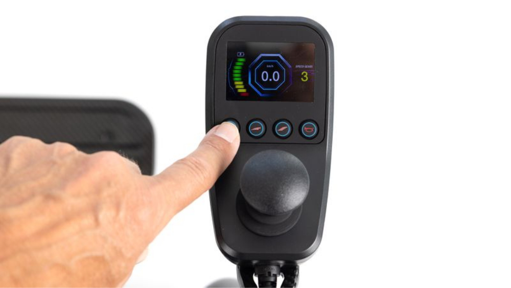 A finger presses a button on a Monarch Mobility MM60 Carbon control panel, which features a digital display for speed and gear settings above a joystick.