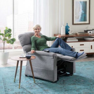 A woman relaxes with her legs elevated, smiling, in a gray Comforter with MaxiComfort Lift Chair. Beside her, a side table displays a blue mug and a book. The modern room features a large window and a blue rug.