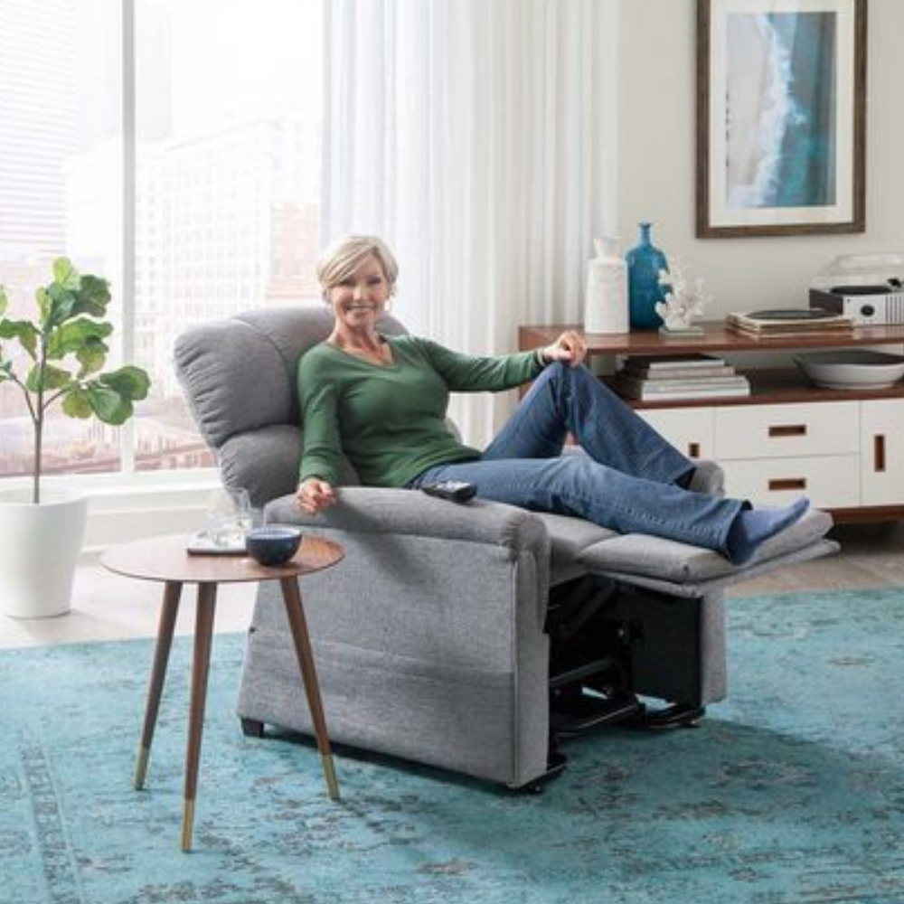 A woman relaxes with her legs elevated, smiling, in a gray Comforter with MaxiComfort Lift Chair. Beside her, a side table displays a blue mug and a book. The modern room features a large window and a blue rug.