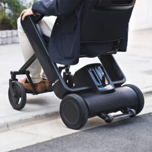 A person using the WHILL Model F, a black electric wheelchair, navigating a curb outdoors with emphasis on the wheelchair’s wheels and mechanism.
