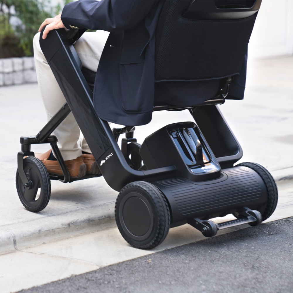 A person using the WHILL Model F, a black electric wheelchair, navigating a curb outdoors with emphasis on the wheelchair’s wheels and mechanism.