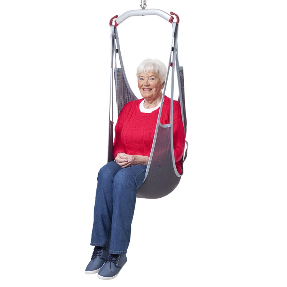 An elderly woman with white hair, in a red sweater and blue jeans, sits suspended from above in a gray Molift EvoSling Polyester Mediumback lifting sling seat.