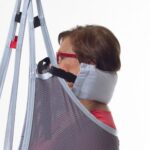 An older adult with short hair and red glasses sits in a Molift EvoSling HeadSupport mesh sling lift, supported by padded straps around the neck and head.