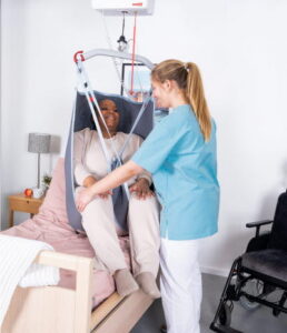A caregiver assists a seated patient in an Rgo Highback Padded sling lift next to a bed in a medical or care facility room.