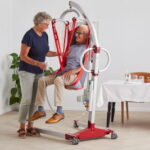 An older man sits in a red patient lift with a Molift Rgo Highback Net while a woman operates the device in a home setting, with a dining table and plants visible in the background.