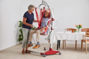An older man sits in a red patient lift with a Molift Rgo Highback Net while a woman operates the device in a home setting, with a dining table and plants visible in the background.