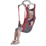 An elderly woman sits in a Molift RgoSling Comfort Highback Net gray and red mesh sling lift, wearing slippers and casual clothes, with her hands resting on her lap.