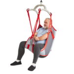 An older man sits in a red and gray Rgo Mediumback Net patient lift sling, suspended off the ground with his arms crossed, wearing a light blue shirt, dark pants, and black slippers.
