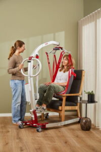 A woman uses a mobile patient lift to assist another woman seated in an Rgo Highback Padded sling on a chair in a living room.