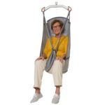 An older woman, wearing glasses, a yellow top, cream pants, and gray shoes, smiles at the camera while seated in a gray Molift UnoSling HighBack sling suspended by a metal frame.