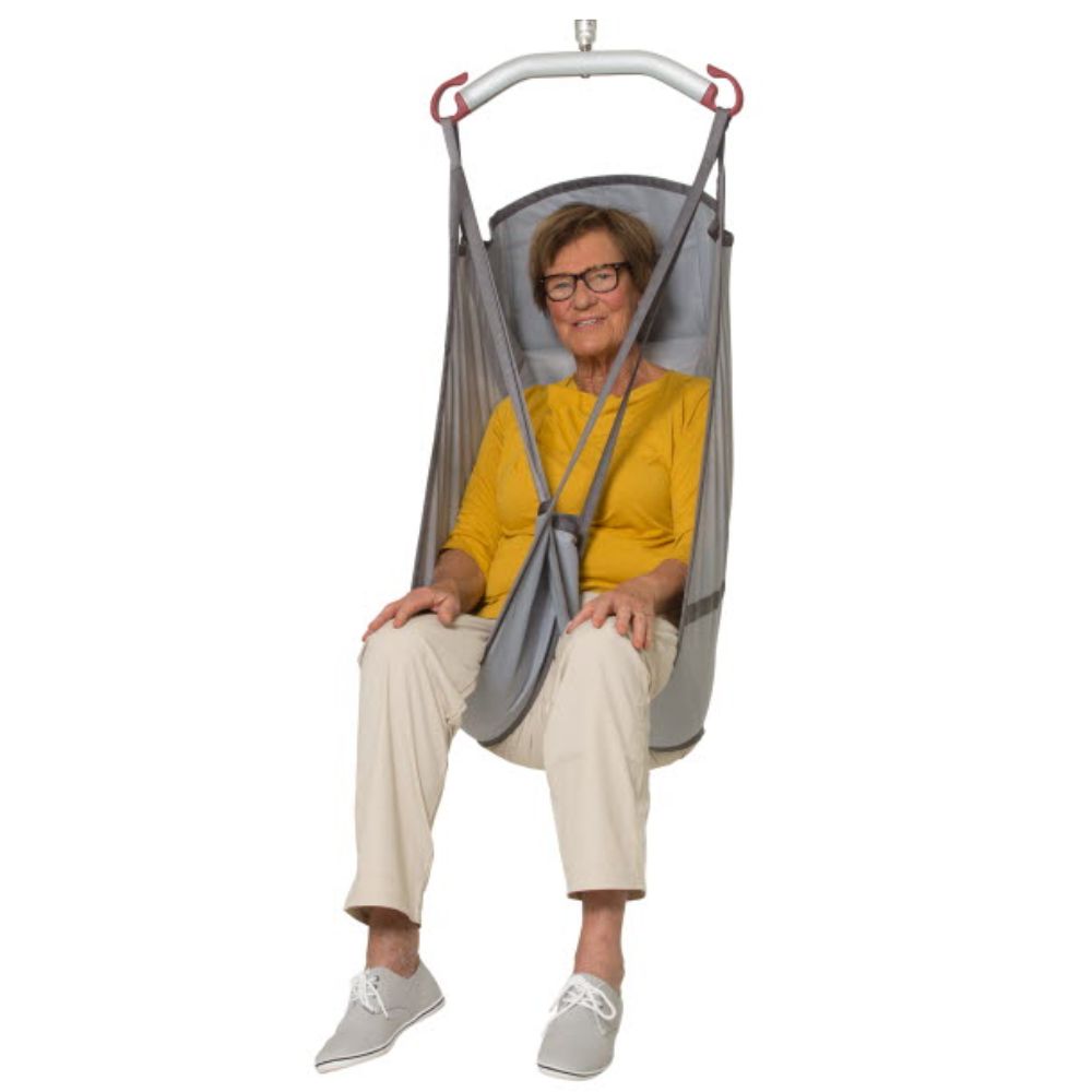 An older woman, wearing glasses, a yellow top, cream pants, and gray shoes, smiles at the camera while seated in a gray Molift UnoSling HighBack sling suspended by a metal frame.