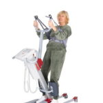 A woman in a green tracksuit stands on an Rgo Standup medical lift, holding the handles and supported by a harness.