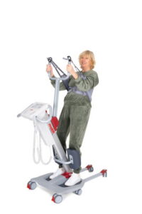 A woman in a green tracksuit stands on an Rgo Standup medical lift, holding the handles and supported by a harness.