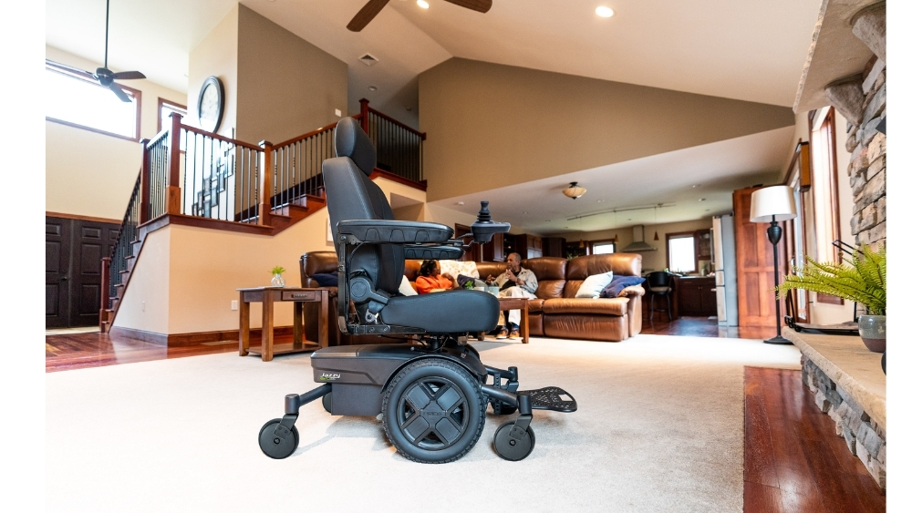 A black Jazzy EVO 613 Li Power Wheelchair sits on a carpet in a spacious living room, with two people talking on a couch in the background.