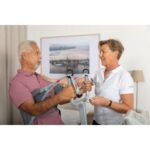 A healthcare worker helps an older man use a Nova Stand-Up lift in a bright room with a framed picture on the wall.
