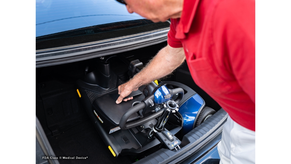 A person in a red shirt points to a compact Go Go Elite Traveller 3-Wheel Scooter stored in the trunk of a car.