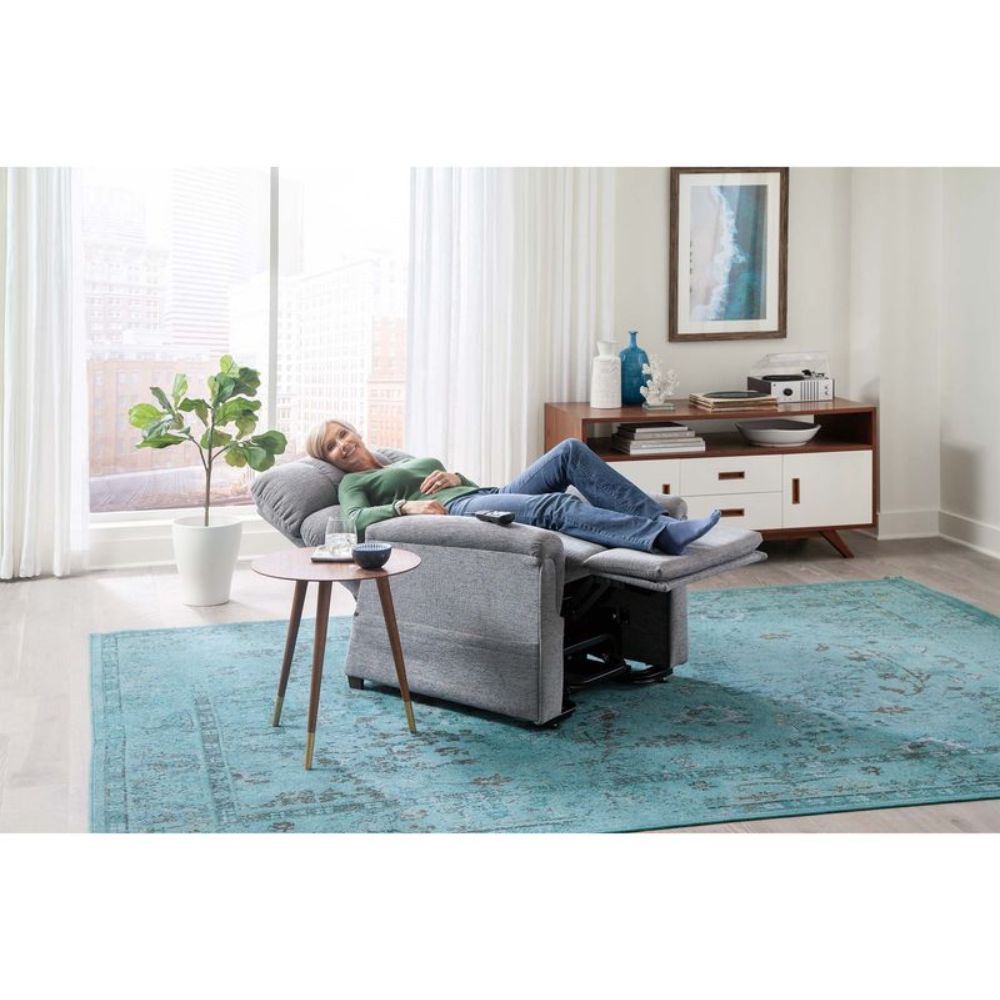 A woman relaxes in a Comforter with MaxiComfort Lift Chair in a modern living room with a teal rug, side table, potted plant, and console by large windows.