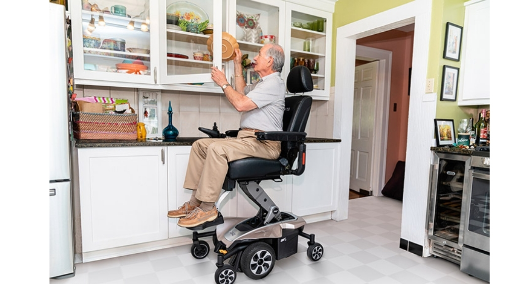 An older man in a Jazzy Air® 2 Power Wheelchair is reaching into a kitchen cabinet to put away a dish, with the wheelchair’s seat elevated to help him access the higher shelf.