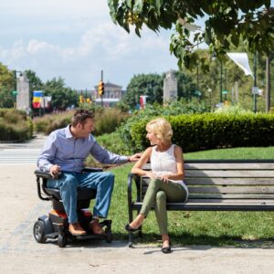 A man in a Go Chair Power Wheelchair and a woman sitting on a bench are talking in a park on a sunny day.