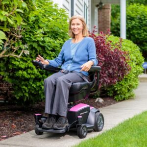 A woman sits in a Go Chair Power Wheelchair on a sidewalk next to a green shrub and red bush, wearing a blue cardigan and gray pants, outside a building.