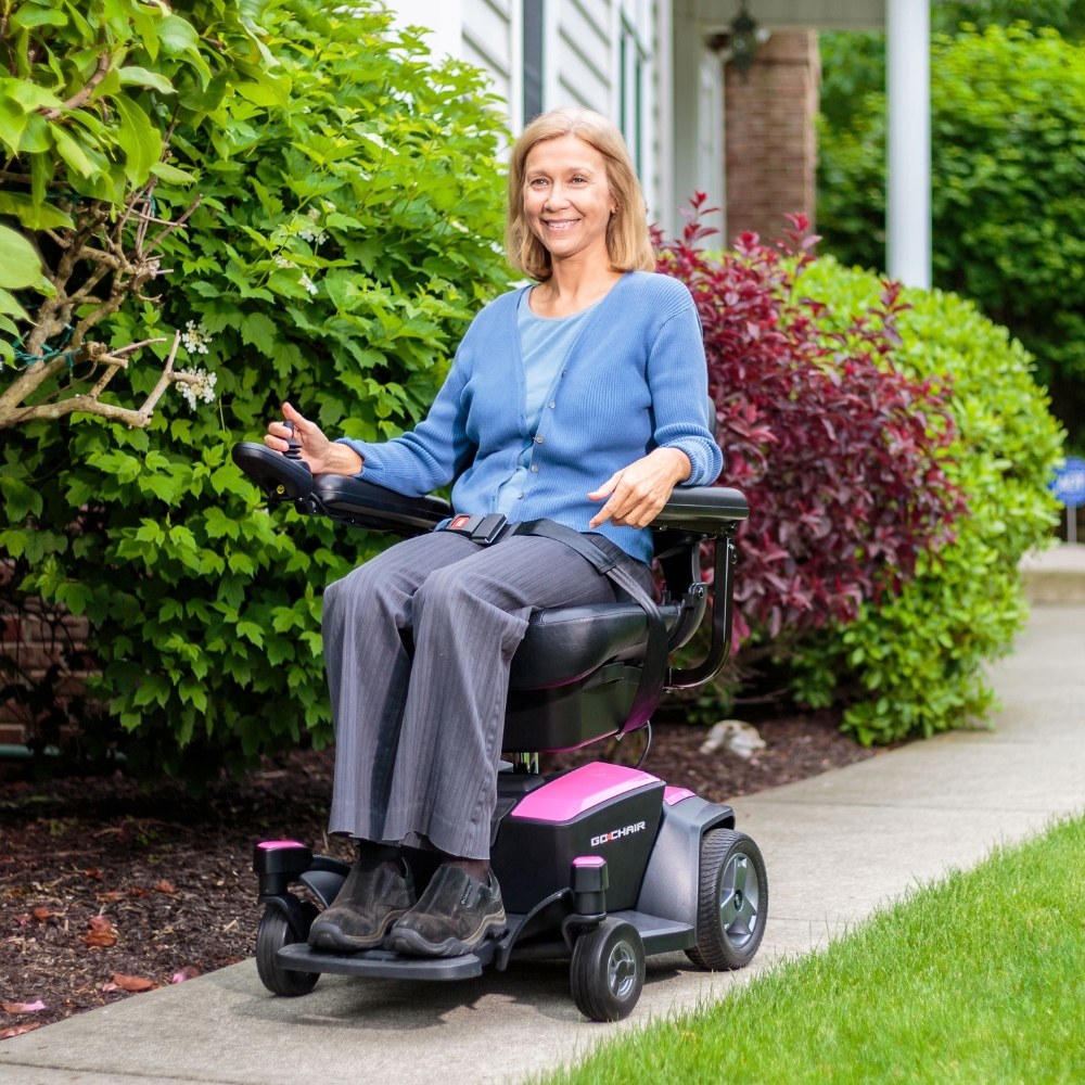 A woman sits in a Go Chair Power Wheelchair on a sidewalk next to a green shrub and red bush, wearing a blue cardigan and gray pants, outside a building.
