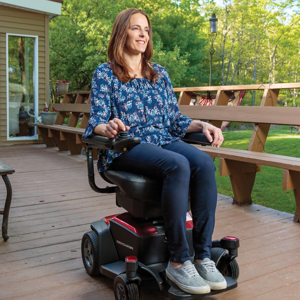 A woman sits on a wooden deck in a Go Chair Power Wheelchair, wearing a blue floral blouse and dark pants, with green trees and a house in the background.