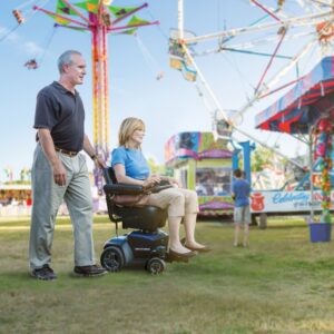 A man pushes a woman in a Go Chair Power Wheelchair at an outdoor carnival, with a Ferris wheel and colorful booths in the background.