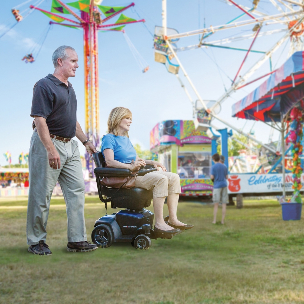 A man pushes a woman in a Go Chair Power Wheelchair at an outdoor carnival, with a Ferris wheel and colorful booths in the background.