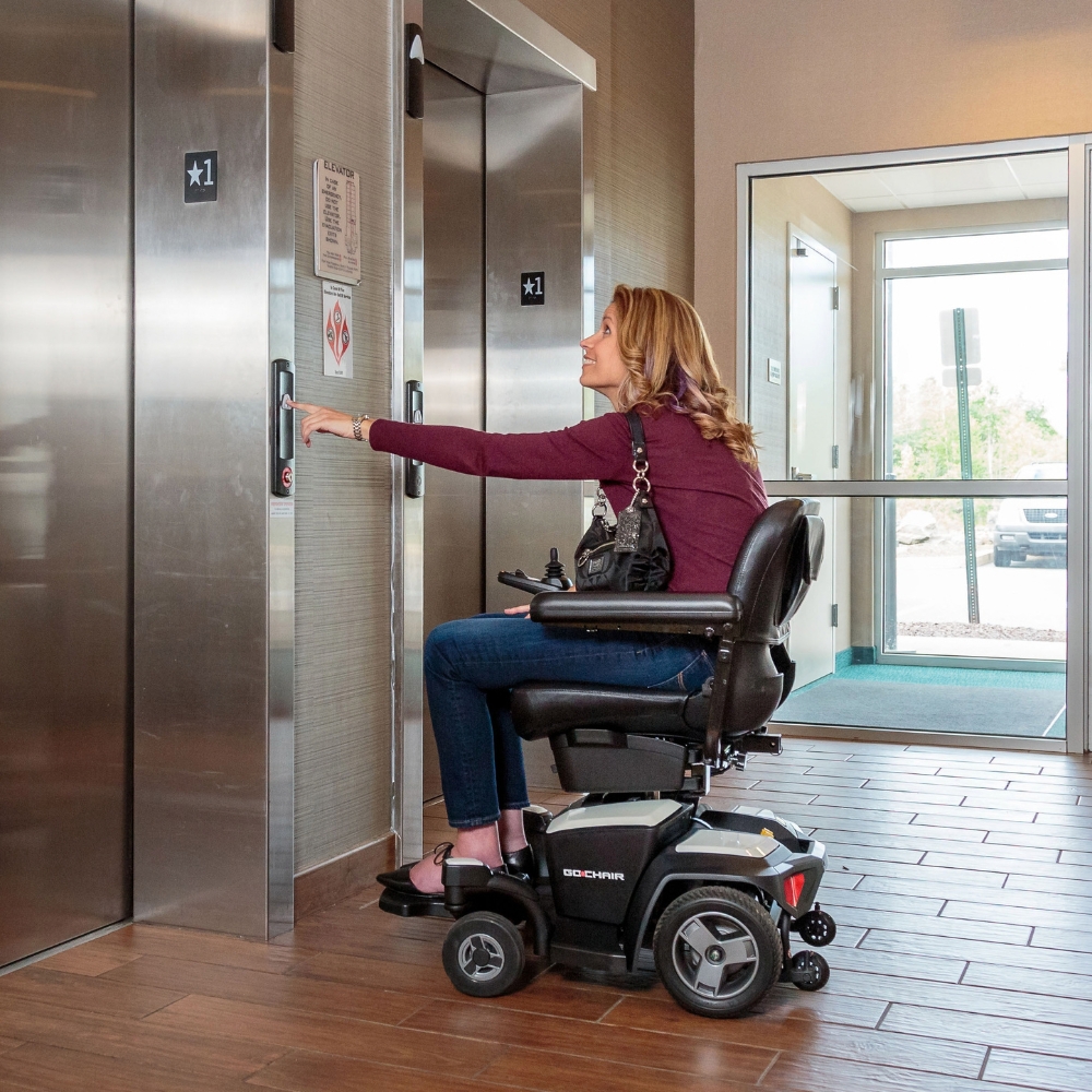 A woman in a Go Chair Power Wheelchair presses the elevator button in a building lobby with tile flooring and natural light.