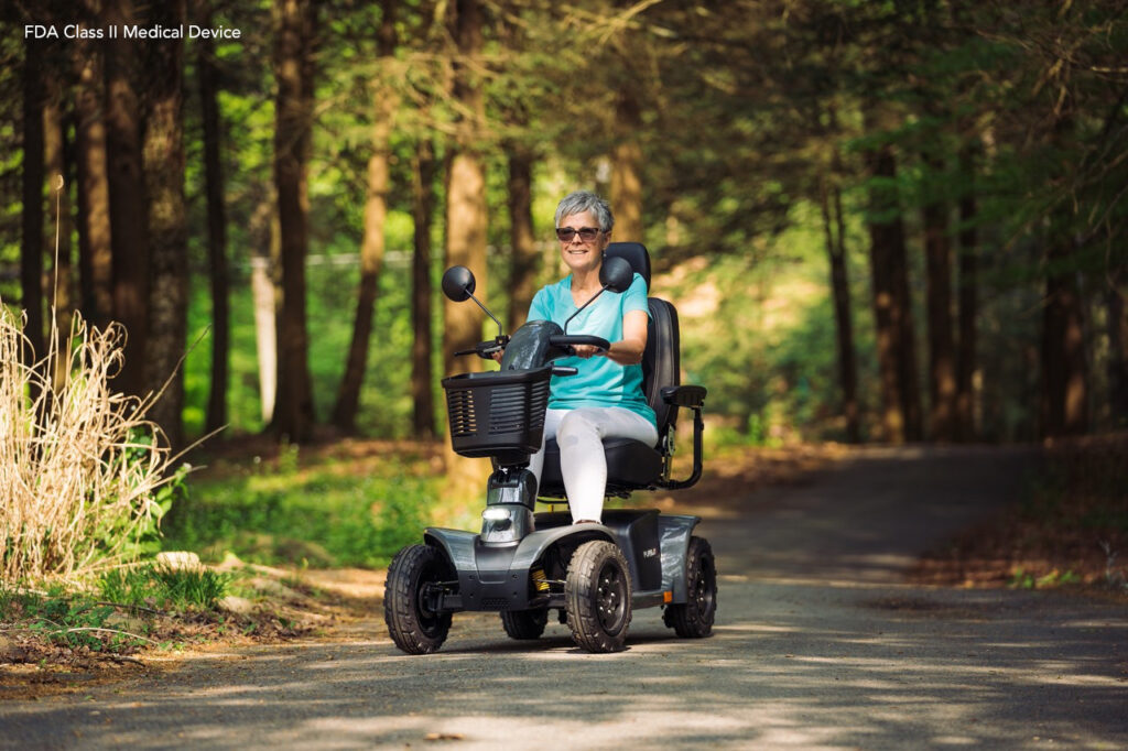 An older woman wearing sunglasses and light blue clothing rides a mobility scooter on a paved path through a wooded area on a sunny day.