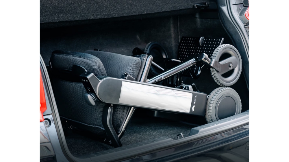 The WHILL Model F electric wheelchair is shown folded and placed in the trunk of a car, demonstrating how it fits compactly in the cargo space.