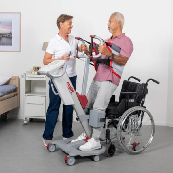 A caregiver skillfully uses the Molift Quick Raiser 205 to help an elderly man transition from his wheelchair in a room with a cozy bed, small table, and framed picture, creating a comforting setting for this important routine.