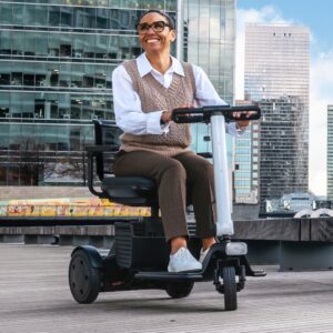A person in glasses and business attire sits outdoors on a WHILL Model R 3-Wheel scooter, with tall modern buildings visible in the background.