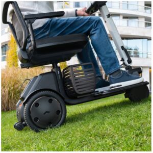A person sits on a WHILL Model R 3-Wheel mobility scooter outdoors on grass, with contemporary apartment buildings and landscaped greenery in the background.