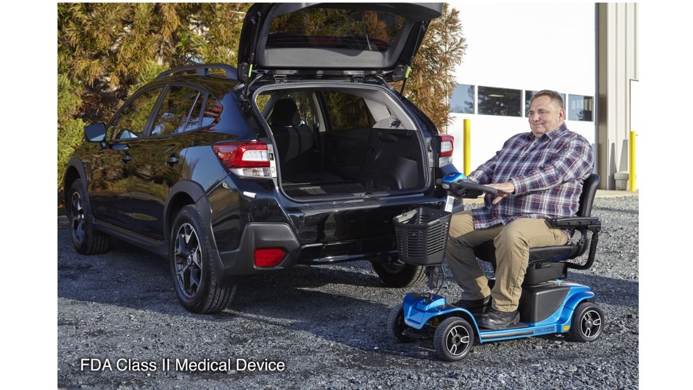 A man sits on a blue Revo® 2.0 4-Wheel Scooter beside the open trunk of an SUV in a parking lot. Text in the corner reads, “FDA Class II Medical Device.”.