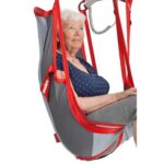 Elderly woman seated in a Molift EvoSling HighBack Padded, gray and red mobility sling lift, wearing a navy polka dot shirt and gray shorts, looking slightly upward.