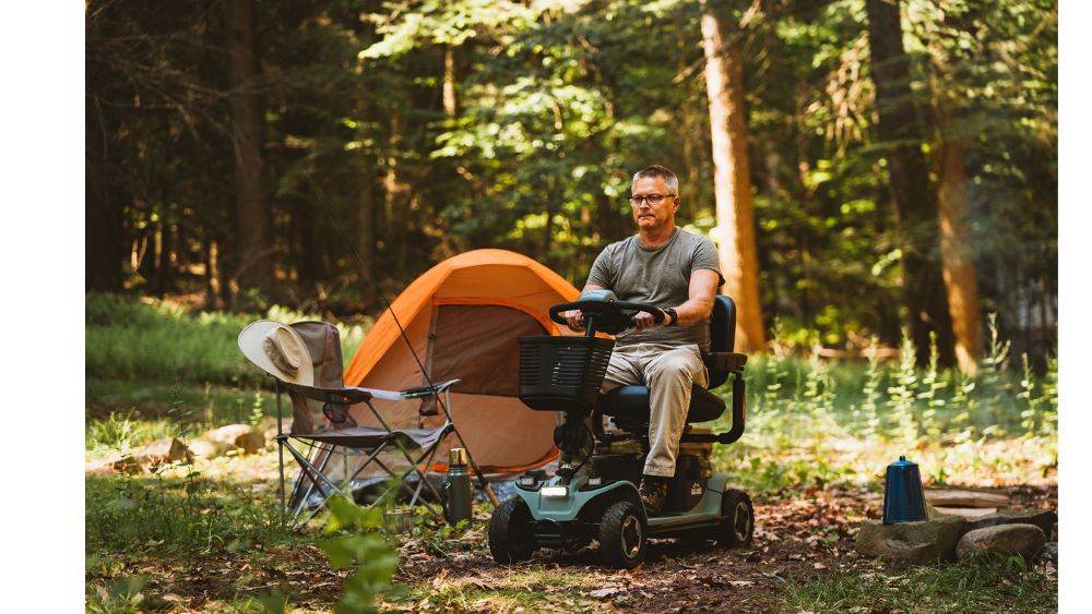 A man rides a Baja™ Bandit 4-Wheel Scooter near an orange tent and a camping chair at a forested campsite during daytime.