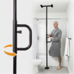 A man in a gray robe holds the Security Pole, featuring a rotating handle, which is installed as a floor-to-ceiling support beside a bathtub in a white bathroom.