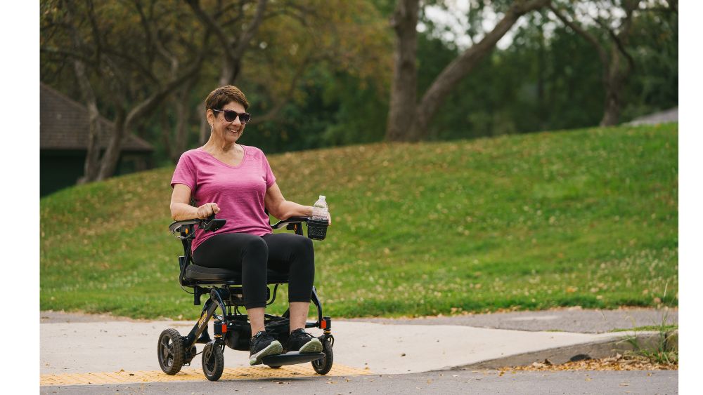 A woman wearing sunglasses and a pink shirt sits in a Jazzy Carbon 27 Power Wheelchair on a paved path, holding a water bottle, with grass and trees in the background.