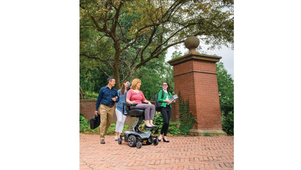 Four adults, including one woman using a Jazzy Air® 2 Power Wheelchair, walk and talk together on a brick path surrounded by trees and a brick pillar.