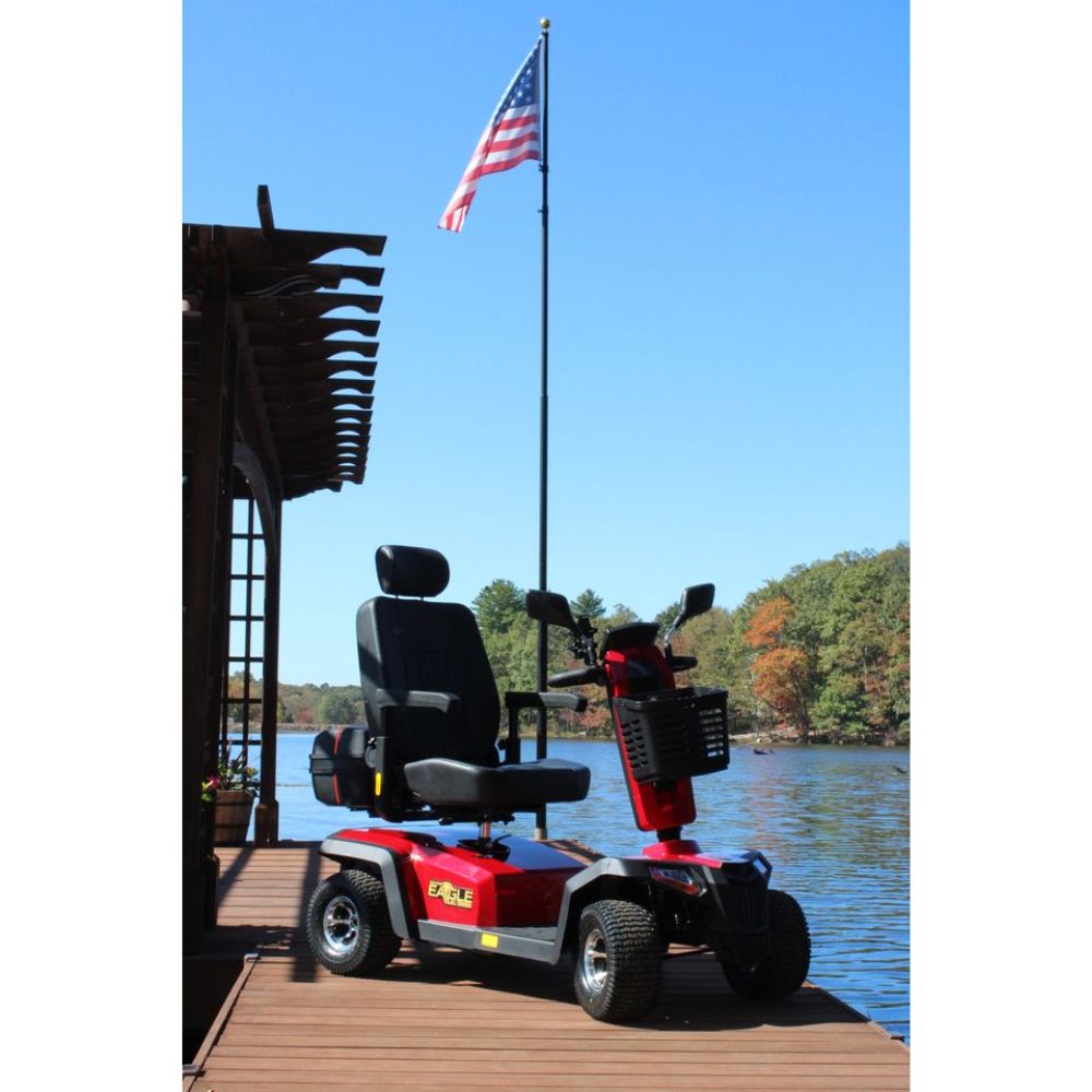 An Eagle All-Terrain Scooter in vibrant red is parked on a wooden dock near a lake, with an American flag displayed on a tall pole in the background.