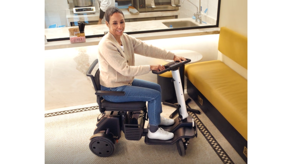 A woman sits on a WHILL Model R electric mobility scooter indoors, smiling at the camera near a yellow cushioned bench and a countertop.