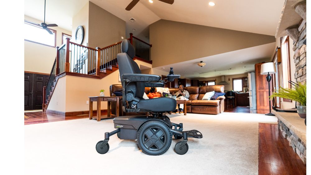 A Jazzy EVO 613 Power Wheelchair sits on a carpet in a spacious living room with a staircase, sofa, and people seated in the background.