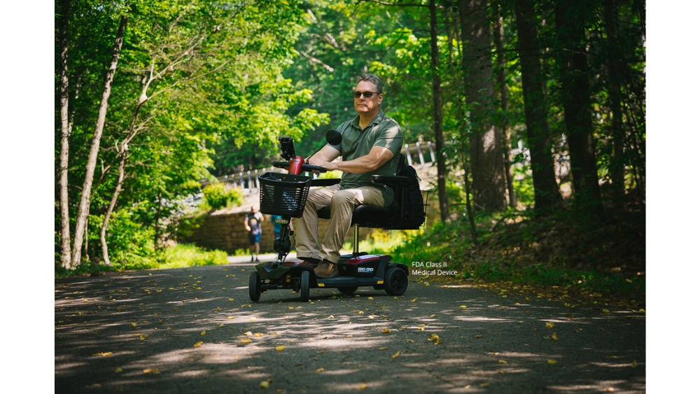 A man rides a Go Go® Endurance AL+ 4-Wheel Scooter along a paved path in a wooded park area on a sunny day.