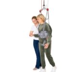 A therapist supports a woman walking with the UnoSling Ambulation Vest, suspended from the ceiling—an effective aid for rehabilitation and physical therapy.