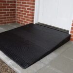The Approach Rubber Threshold Ramp, in black, leads to a white door surrounded by red brick walls, all set upon gray and concrete tiles.