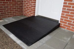 The Approach Rubber Threshold Ramp, in black, leads to a white door surrounded by red brick walls, all set upon gray and concrete tiles.