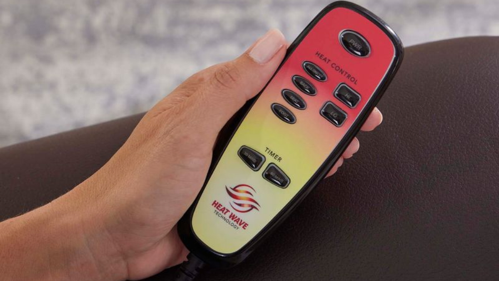 A hand holds a Heat Wave Technologies remote control—with heat control, timer, and power buttons—resting against a dark cushioned surface, making it the perfect accessory for relaxing in your Cloud Lift Chair w/ MaxiComfort.