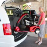 A woman stands by a white vehicle as a red mobility scooter is lifted into the cargo area using a Bruno VSL-6000 Curb-Sider® Hoist Style Interior Lift.