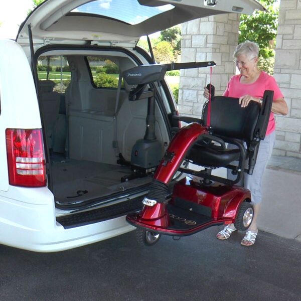 An older woman uses a Bruno VSL-6000 Curb-Sider® Hoist Style Interior Lift to load a red mobility scooter into the back of a white minivan outside a building.